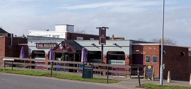 Single-storey modern pub of red brick and grey cladding with a small pitched roof porch, a maroon fascia reading The Reiver, closed purple parasols and boards reading Food Served, seen behind a timber rail fence and grass verge with a matching pole sign