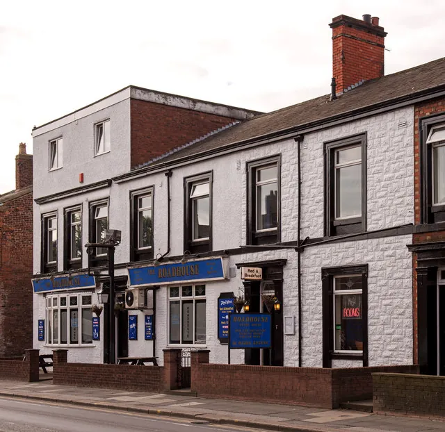 Two-storey white painted pub with black window frames, blue fascias reading The Roadhouse, a Bed & Breakfast sign over the door and a lit Rooms sign in a window, behind a low brick wall on a terraced street