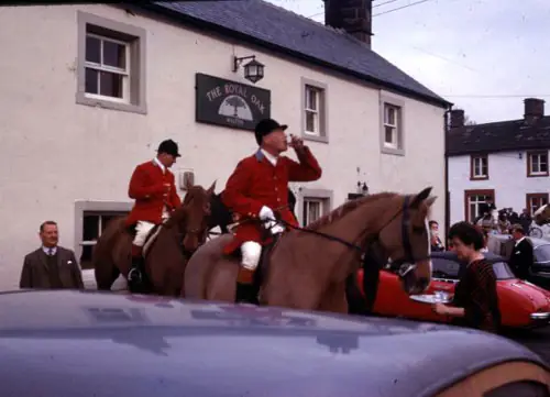 Two huntsmen in red coats and black caps on horseback outside a whitewashed two-storey inn, one drinking from a glass, with a hanging sign reading The Royal Oak, Welton, showing an oak tree; a man in a suit stands to the left, a woman holds a tray beside the horses, and a red sports car is parked behind