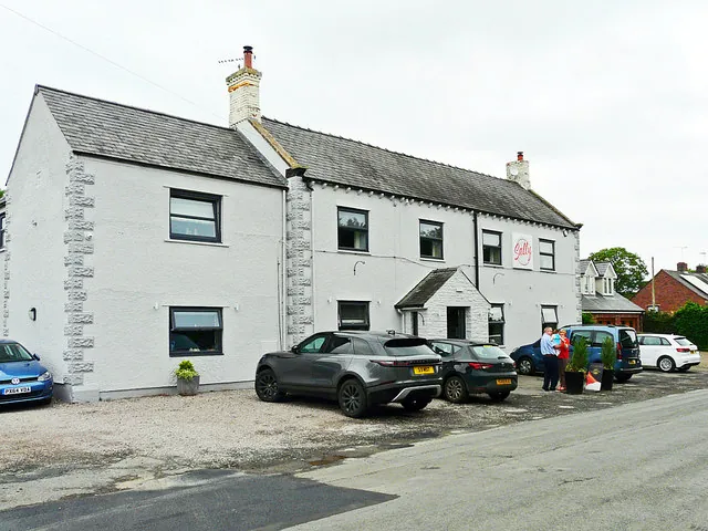 Two-storey white rendered inn with grey stone quoins, a slate roof and a small gabled porch, a round red sign on the front, and cars parked on the gravel forecourt