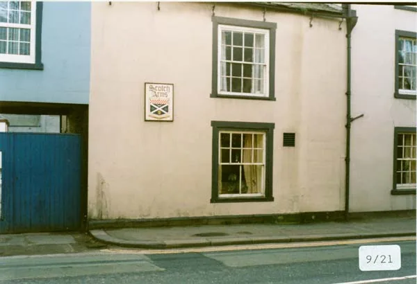 Colour photograph of the end of a rendered building with a painted sign reading Scotch Arms and a saltire, a blue wooden gate beside it and the pavement in front