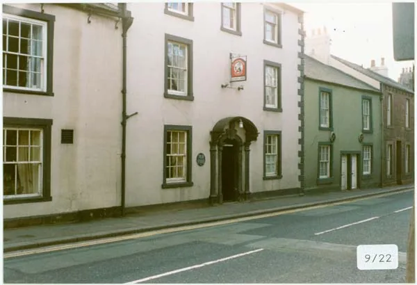 Colour photograph of a rendered three-storey frontage with a round-arched stone doorway, sash windows, a small hanging sign and a plaque beside the door, on a quiet street