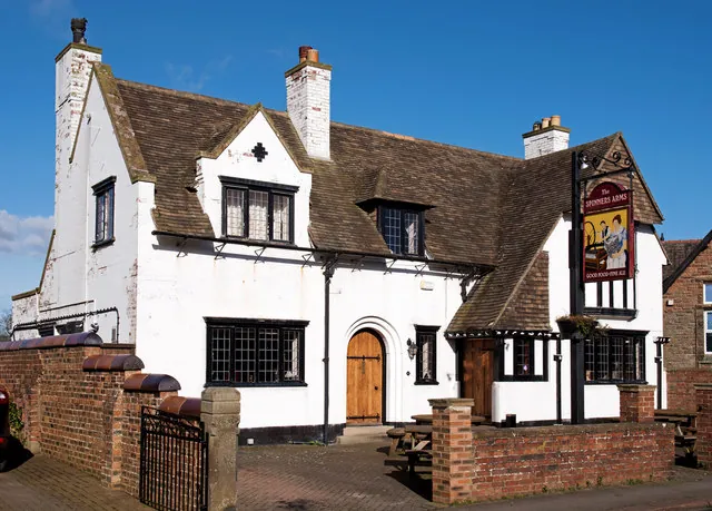Whitewashed two-storey pub with a steep brown tiled roof, tall chimneys, mock Tudor gables and black leaded windows, with an arched oak front door. A tall post sign reading The Spinners Arms stands at one end behind a low red brick wall, with picnic benches on the forecourt.