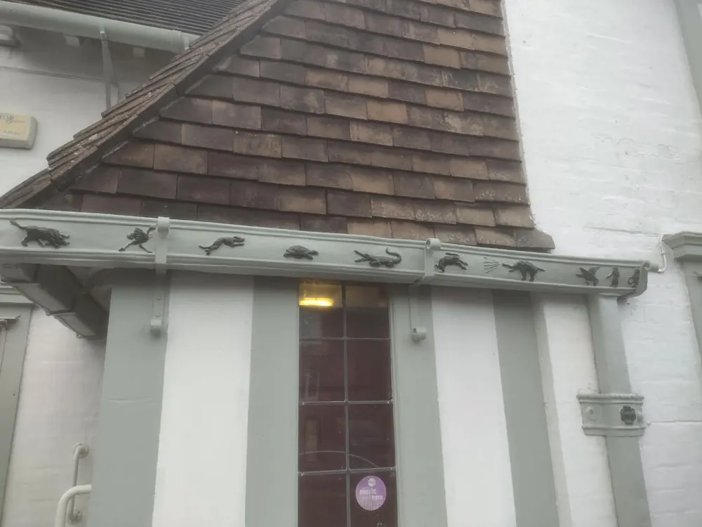 Pale green cast-metal gutter running along the front of a white-rendered building under a tiled gable, with small dark figures set along it: a boar or bull, a hound, a tortoise, a crocodile, a hare, an elephant, a winged beast and two or three tiny human-like figures
