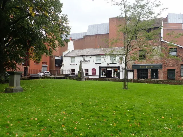 Small two-storey whitewashed pub with a slate roof and black window surrounds, lettered The Sportsman, seen across a wide grassy churchyard with an old stone monument and young trees, between taller red brick buildings including a shop called Salon Panache