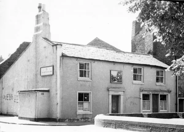 Black and white photograph of a low two-storey rendered pub with a stepped gable chimney, a sign reading The Sportsman Inn on the gable end, a picture sign between the upper windows and chalked names on the wall