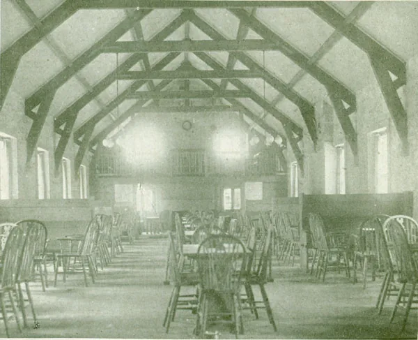 Old printed photograph of a long hall with an open timber roof of king-post trusses, rows of tables with wheelback chairs, a clock and a gallery at the far end