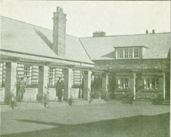 Old printed photograph of a long low stone building with a slate roof, a tall chimney and a covered walkway on square pillars, with two men standing under it and a lawn in front
