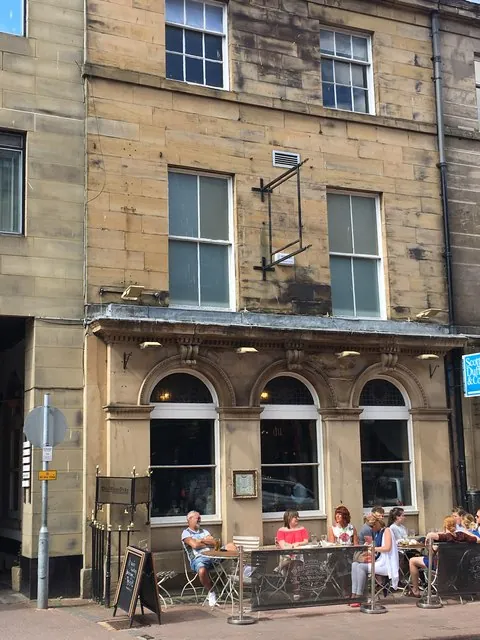 Tall sandstone town building with large sash windows on the upper floors and a ground floor of three round arched windows framed by carved pilasters under a small canopy, with an empty sign bracket on the wall and people sitting at pavement tables behind barriers