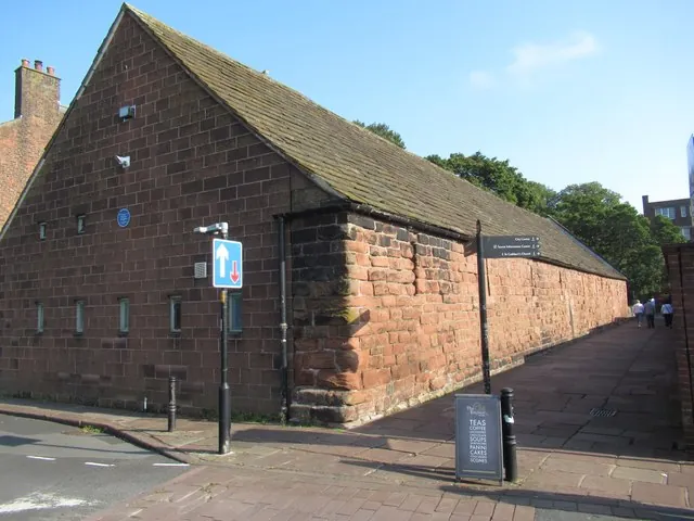Long red sandstone barn with a steep stone slab roof, small slit windows and a blue plaque on the gable end, a paved path running along its side wall, and a pavement board out front advertising teas, coffee, soups, panini, cakes and scones