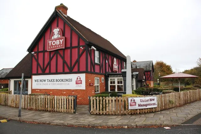 Red brick roadside restaurant pub with a tall mock Tudor gable painted dark pink and black, and a Toby Carvery sign with a figure in a tricorn hat near the apex. A banner reads We Now Take Bookings, and a low picket fence carries an Under New Management sign.
