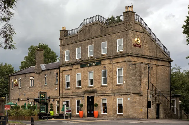 Tall three-storey sandstone building with iron railings around a flat roof terrace, a green sign reading The Turf above the central doorway flanked by orange planters, a lower two-storey stone wing to the left and an external iron staircase on the right