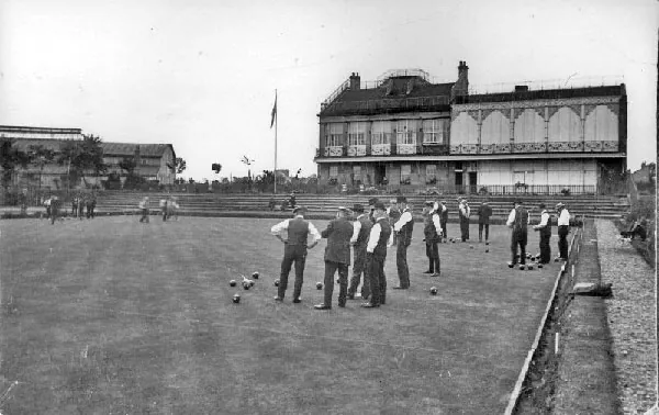 Black and white photograph of men in waistcoats and shirtsleeves playing bowls on a large green, with a flagpole and a tall building with an ornate iron balcony and covered grandstand front behind