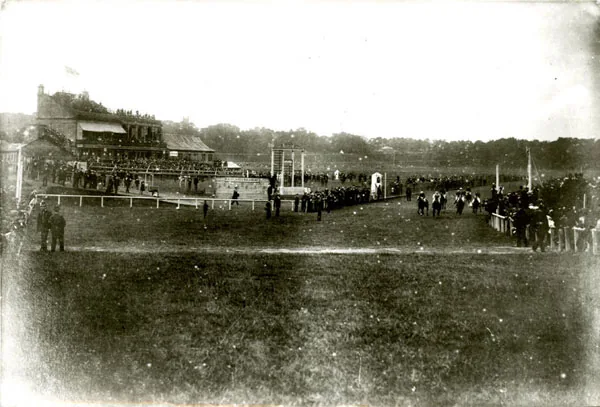 Old photograph of a racecourse with crowds along the rails and in a grandstand on the left, a judge's box and posts by the track and a man in the foreground