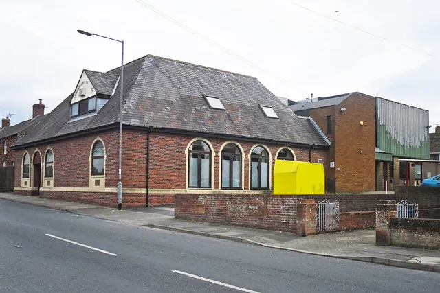 Single-storey red brick building with a hipped slate roof, a row of round-arched windows with cream surrounds, a small dormer and roof lights, a yellow bin by the wall and a taller modern brick extension behind