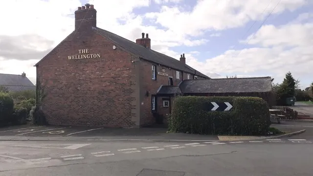 Two-storey red brick pub with a slate roof and tall chimney, The Wellington lettered in gold on the gable end and again along the side, with a lower outbuilding and a clipped hedge carrying a chevron road sign at the corner