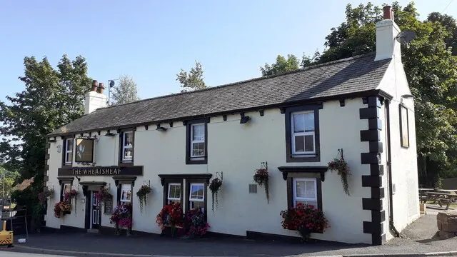 Cream rendered two-storey pub with a grey slate roof, black window surrounds, black and white quoins at the corner, a sign reading The Wheatsheaf over the door and colourful hanging baskets and window boxes, with tall trees behind under a clear blue sky