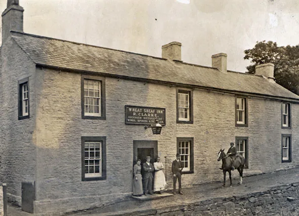 Old sepia photograph of a long two-storey stone inn with a board over the door reading Wheat Sheaf Inn, R. Clarke, licensed retailer of wines and spirits, a group of people at the door and a man on horseback alongside